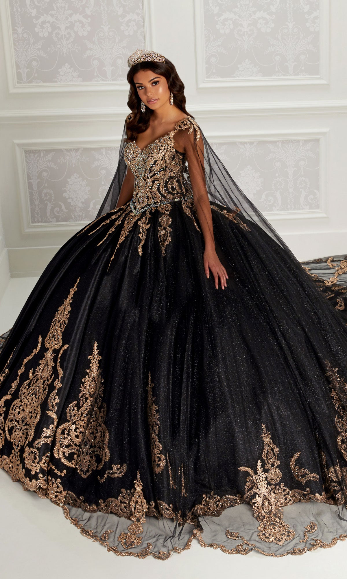 A woman in the Quinceanera Dress PR22141 By Princesa, a black ball gown with gold embroidery and a corset back, poses indoors with a tiara against a white paneled wall.